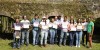 Course participants stand together on grass for a photo in front of a moss-covered bridge, proudly presenting their course completion certificates and smiling. thumbnail